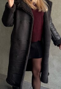 Μαύρο δερμάτινο παλτό με αφράτη, σκούρα καφέ επένδυση; διαθέτει μεγάλες κουμπώματος, φαρδύ γιακά και έχει μήκος μέχρι το γόνατο. Φοριέται πάνω από ένα μπορντό πουλόβερ.