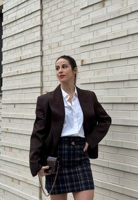 Femme en blouse blanche, blazer sombre et jupe à carreaux tenant un petit sac à main sombre, debout près d'un mur en briques claires, regardant vers la gauche.
