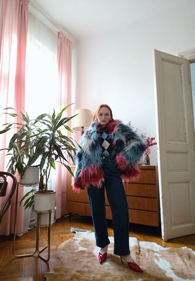 Mujer con un abrigo peludo de colores, suéter de rombos, vaqueros oscuros y tacones rojos, de pie sobre una alfombra de piel de vaca en una habitación luminosa con plantas.