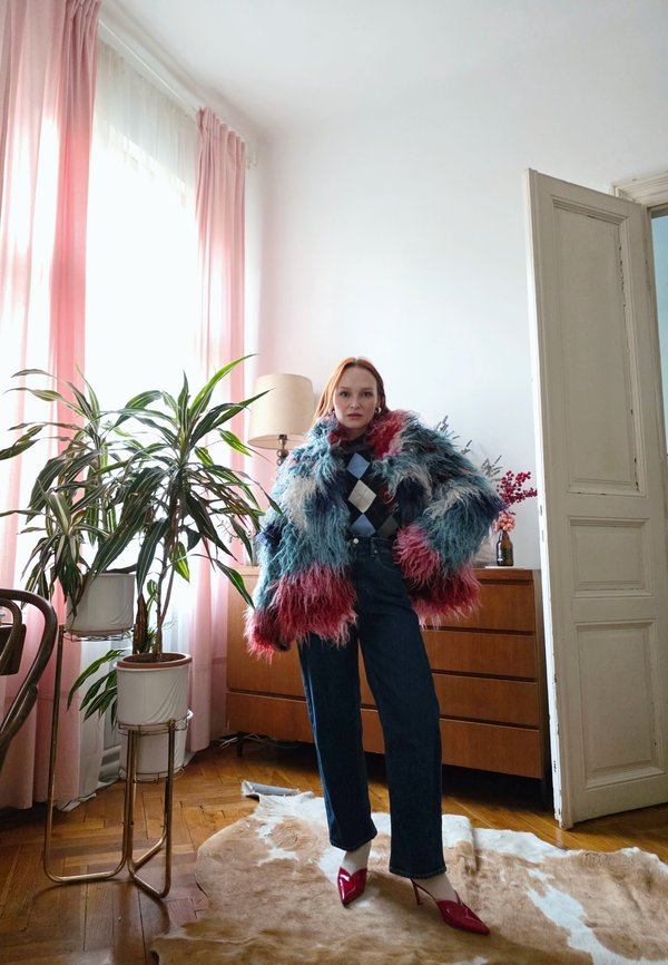 Woman wearing a colourful shaggy coat, argyle jumper, dark jeans, and red heels stands on a cowhide rug in a bright room with plants.