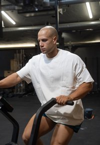 Bærer på en sort træningsmaskine, iført en løs hvid bomulds skjorte og teal shorts, med definerede muskler og et fokuseret udtryk.