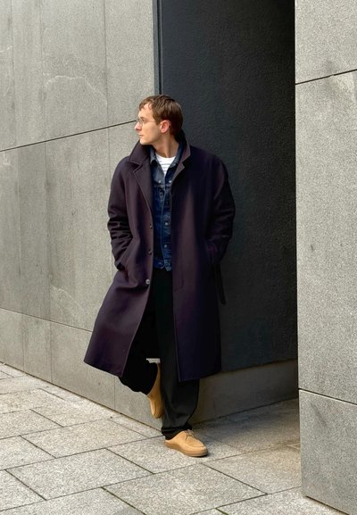 Largo abrigo negro sobre una chaqueta de denim, combinado con pantalones negros y zapatos marrones. Fondo de pared de piedra gris. Pose de frente, de pie.