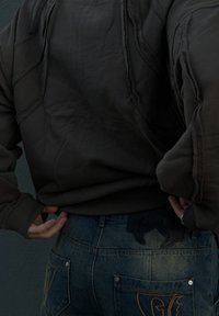 Sweat-shirt marron foncé avec des détails de couture visibles, associé à un jean en denim bleu avec des coutures décoratives et un patch noir.