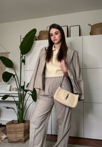 Femme portant un costume à rayures beige et un pull crème, tenant un sac à main beige pâle, debout à l'intérieur près d'une grande plante verte et de placards blancs.