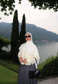 Una mujer con un hijab blanco y un suéter color crema está junto a un lago. Lleva gafas de sol negras y una falda gris con pliegues, sosteniendo un bolso negro.