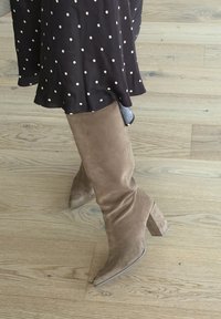 Personne portant des bottes à talons hautes en suède beige jusqu'aux genoux et une jupe foncée à pois blancs, debout sur un sol en bois clair.