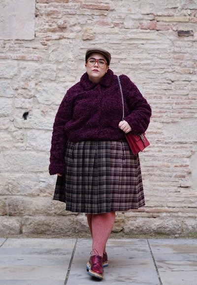 Persona con chaqueta texturizada morada, falda de cuadros, zapatos rojos y sombrero, de pie contra una pared de piedra sosteniendo un bolso rojo con cadena.