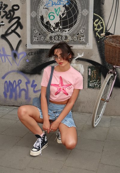 Camisa de manga corta rosa con un diseño de estrella de mar, falda de mezclilla y zapatillas negras de caña alta. El fondo presenta graffiti y una bicicleta.