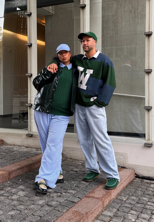 Deux personnes debout sur un sol pavé, l'une portant un pull vert et un pantalon rayé, l'autre un maillot de rugby rayé et un jean clair, toutes deux portant des casquettes.