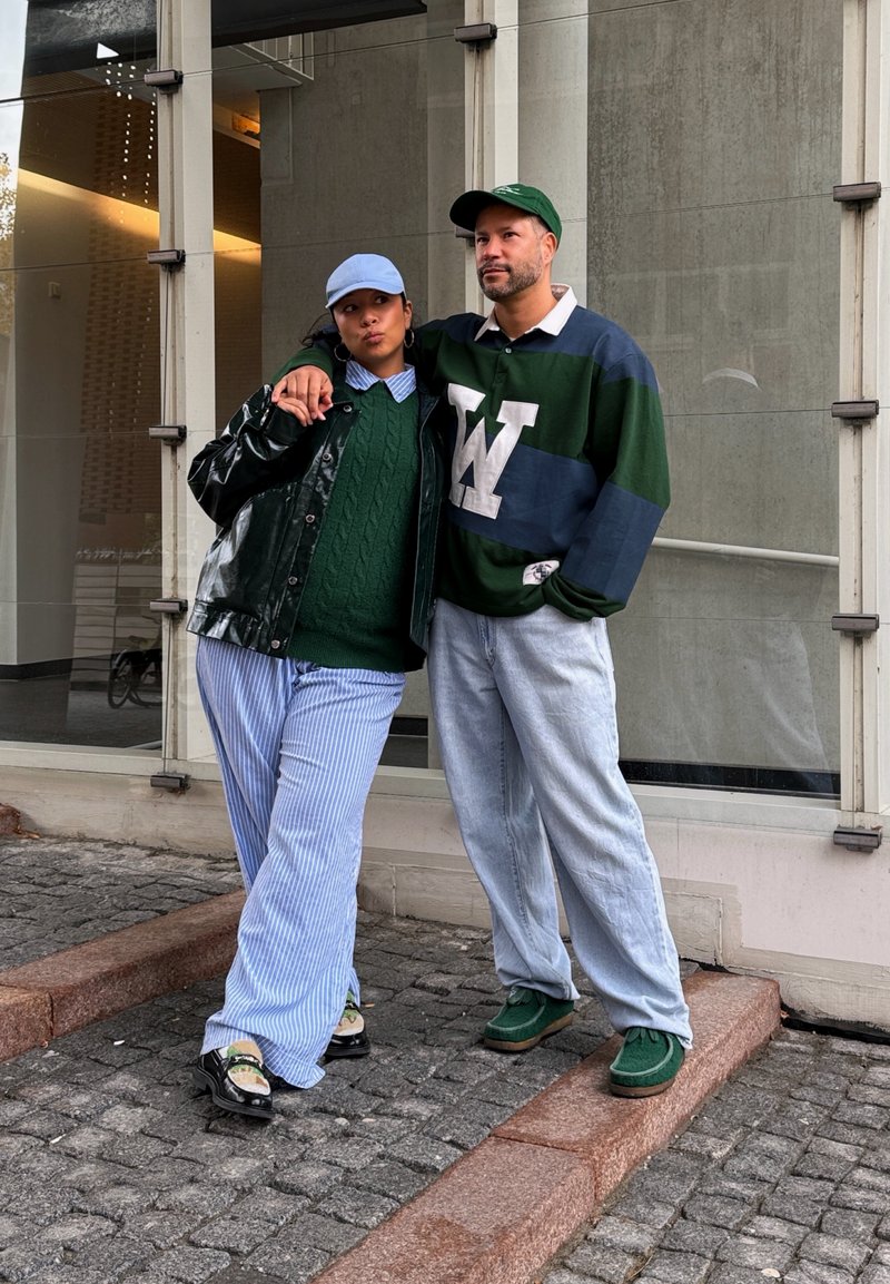 Dvaja ľudia stoja na kamennom dlažobnom koberci, jeden má zelený sveter a pruhované nohavice, druhý pruhované rugby tričko a svetlé rifle, obaja majú na hlave čiapky.