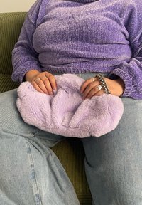 Lilla plysjpute med en teksturert overflate, holdt i hendene. Løse, lyse denimjeans og en lilla strikkegenser er synlige.