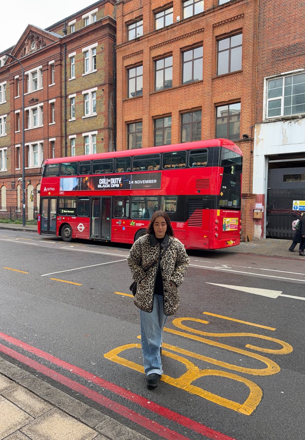 Jeune femme en manteau léopard marchant dans la rue près d'un bus rouge à impériale garé près de bâtiments en briques sous un ciel nuageux.