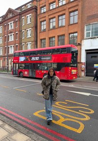 Jeune femme en manteau léopard marchant dans la rue près d'un bus rouge à impériale garé près de bâtiments en briques sous un ciel nuageux.