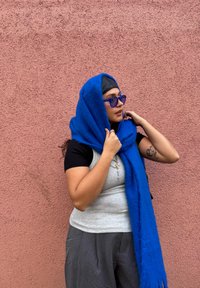 Écharpe bleue et moelleuse drapée sur un haut gris clair. La personne a les cheveux foncés et porte des lunettes de soleil bleues devant un mur rose texturé.