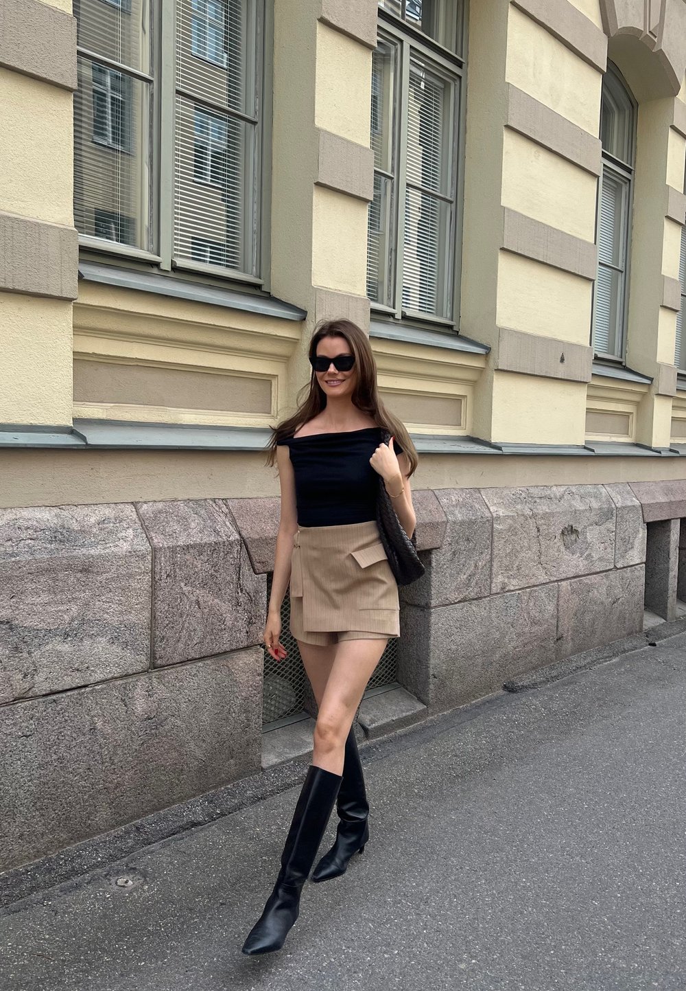 Μαύρη off-shoulder μπλούζα, μπεζ φούστα με πλαϊνές τσέπες, μαύρες μπότες μέχρι το γόνατο. Το μοντέλο στέκεται σε ένα δρόμο δίπλα σε έναν πέτρινο τοίχο.