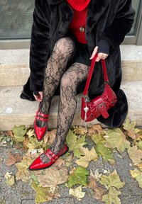 Ballerine in vernice rossa con punta affusolata, con dettagli a fibbia. Collant neri a rete con un motivo e una borsa rossa coordinata. Foglie cadute sullo sfondo.