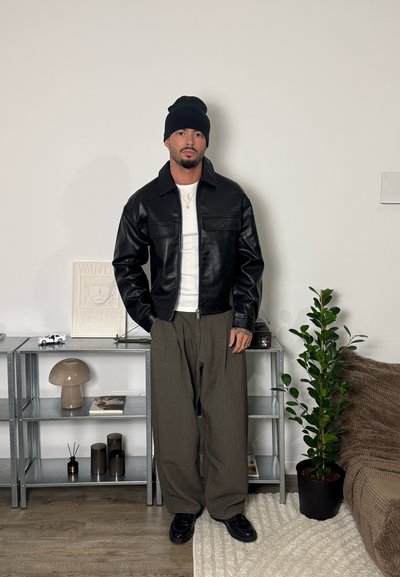 Hombre con gorro negro, chaqueta de cuero negra, camiseta blanca y pantalones marrones holgados, de pie en una habitación minimalista con estanterías de metal y planta en maceta.