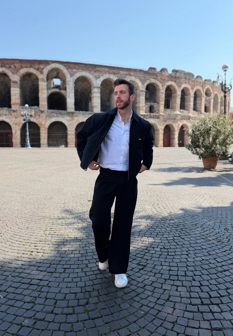 Man wearing black jacket, white shirt, and white shoes walking on cobblestone street near ancient Roman amphitheater under clear blue sky.