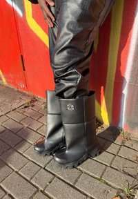 Sorte vandtætte støvler med flad sål, quiltet design, cirkulært logo og en høj, struktureret skaft. Båret med sorte teksturerede bukser.