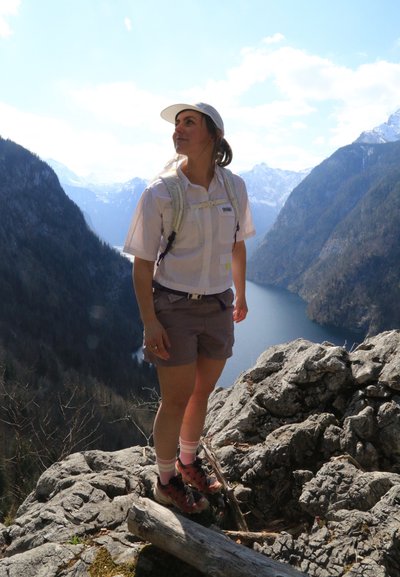 Mujer con gorra blanca y equipo de senderismo de pie en un acantilado rocoso con vistas a un lago y montañas con cumbres nevadas bajo un cielo azul.