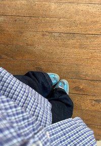 Pantofi slip-on din suede albastru, cu vârful rotund, asortați cu blugi negri largi și o cămașă albastră în carouri, pe o podea din lemn.