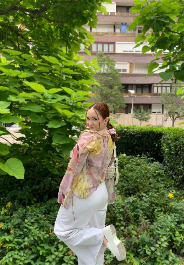 Transparante blouse met bloemenpatroon in pastelkleuren, gecombineerd met witte wijde broek en witte platformschoenen. De achtergrond toont groene bladeren.