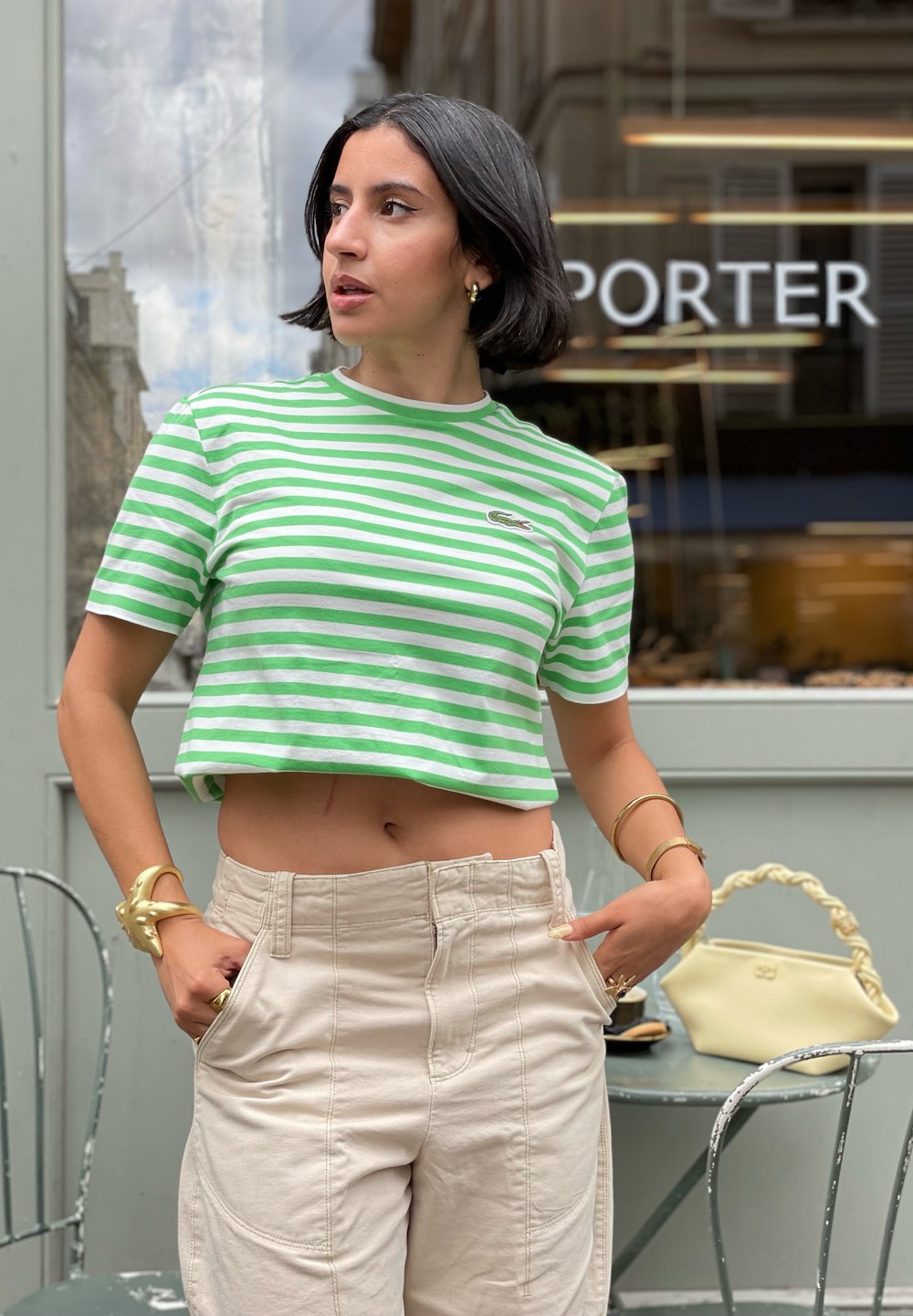 Grün-weiß gestreiftes cropped T-Shirt, kombiniert mit beigen hochgeschnittenen Cargo-Hosen. Auffälliges goldenes Armband als Akzent. Heller Hintergrund.