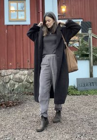Manteau noir surdimensionné, pull gris, et pantalon gris à rayures. Sac à main en cuir marron et bottines marron complètent la tenue. Sol pavé, bâtiment en bois en arrière-plan.