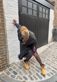 Svart bomberjakke over en denimtopp, svart rysjekjole, prikete strømpebukse og brune ankelstøvler, poserer ved siden av en hvit murvegg og en tredør.