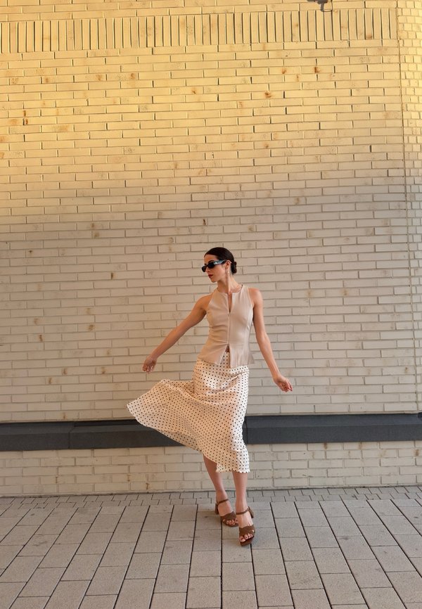 Femme portant des lunettes de soleil, un haut beige sans manches et une jupe blanche à pois, debout sur un pavement carrelé devant un mur en briques clair.