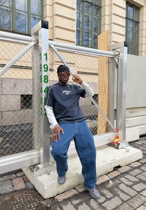 Hombre con gorro oscuro, gafas de sol, camisas superpuestas y jeans sueltos apoyado en una valla metálica de construcción con postes numerados frente a un edificio beige.
