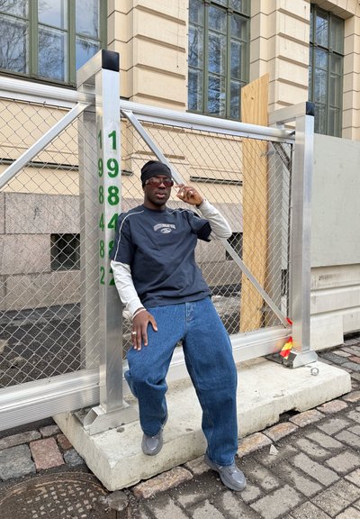 Hombre con gorro oscuro, gafas de sol, camisas superpuestas y jeans sueltos apoyado en una valla metálica de construcción con postes numerados frente a un edificio beige.