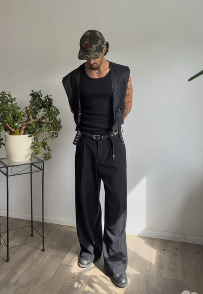 Hombre que lleva una camiseta sin mangas negra, un chaleco negro, pantalones anchos negros, zapatos negros y una gorra de camuflaje, de pie en el interior cerca de una planta sobre un soporte metálico.