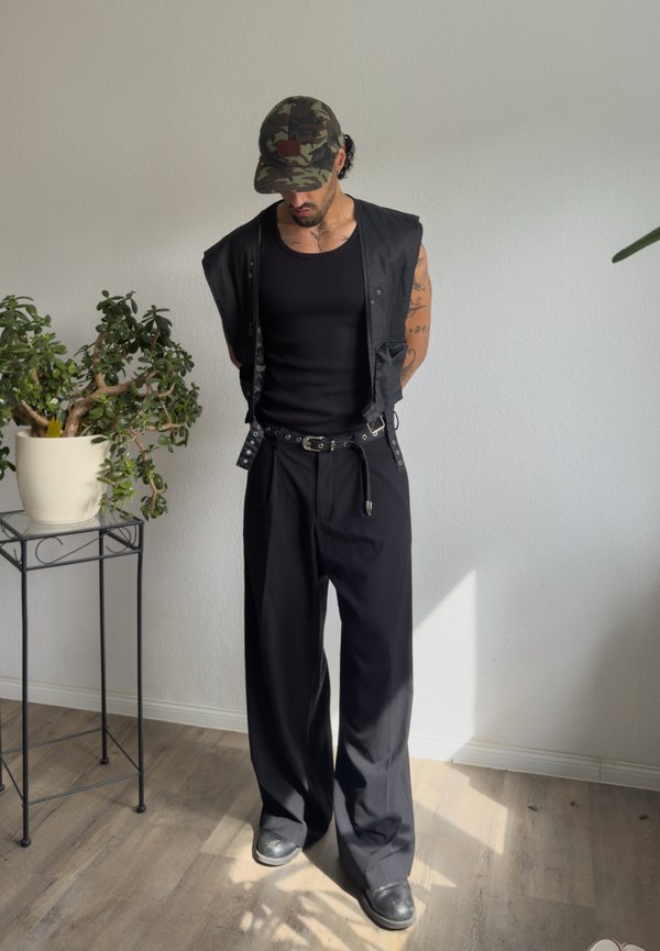 Ein Mann trägt ein schwarzes Tanktop, eine schwarze Weste, weite schwarze Hosen, schwarze Schuhe und eine Camouflage-Kappe und steht drinnen in der Nähe einer Pflanze auf einem Metallständer.