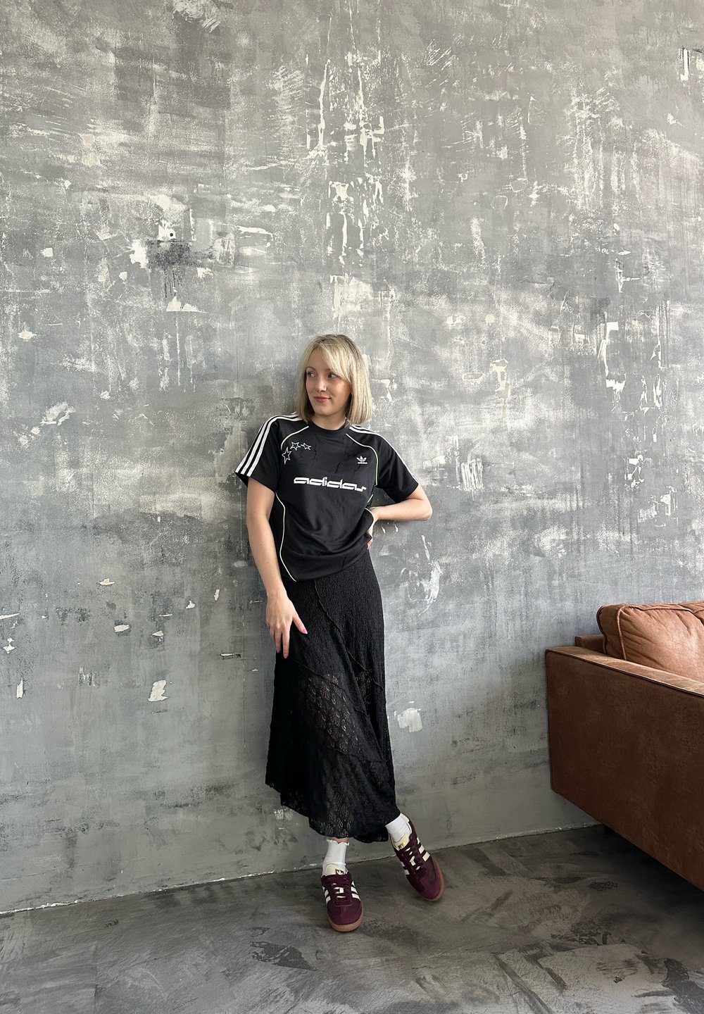 Kvinna i svart Adidas-tröja, svart spetskjol, vita strumpor och vinröda sneakers stående mot en texturerad grå vägg nära en brun soffa.