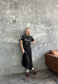 Kvinna i svart Adidas-tröja, svart spetskjol, vita strumpor och vinröda sneakers stående mot en texturerad grå vägg nära en brun soffa.