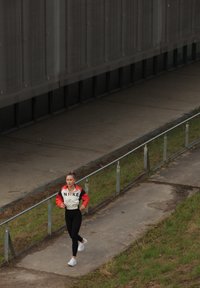 MLADA ŽENA TEKA PO NARAVNEM POTI OB TRAVI, OBLEČENA V ČRNE LEGICE, BELE TENISICE IN RDEČO-BELO JAKNO NIKE.