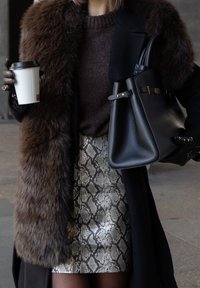 Čierna kabelka so zlatými akcentmi, hnedý golier z umelého kožúška, hnedý pletený sveter, metalická sukňa s hadím vzorom, čierne rukavice, drží hrnček kávy.