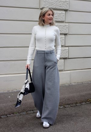 Mujer caminando por la acera, vistiendo un suéter blanco abotonado, pantalones anchos grises, zapatillas blancas, y llevando un bolso negro con una bufanda estampada.