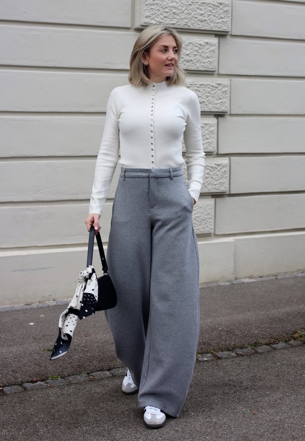 Femeie mergând pe trotuar, purtând un pulover alb cu nasturi, pantaloni gri largi, teniși albi, ținând o geantă neagră cu o eșarfă cu model.