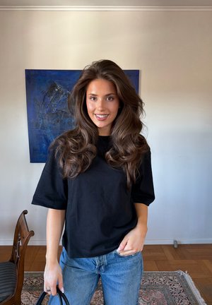 Mujer con cabello largo y ondulado de color castaño lleva camiseta negra y vaqueros azules, de pie en el interior cerca de una silla de madera y una pintura abstracta azul.