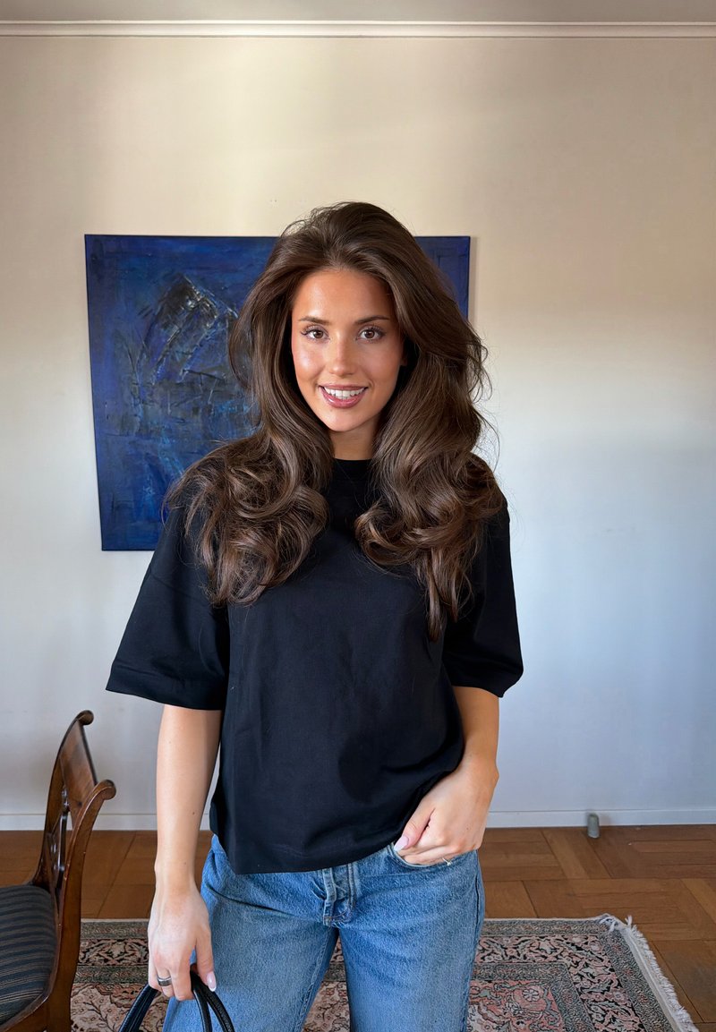 Femme aux longs cheveux bruns ondulés porte un T-shirt noir et un jean bleu, debout à l'intérieur près d'une chaise en bois et d'une peinture abstraite bleue.