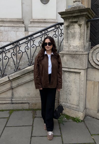 Chaqueta de ante marrón sobre una camisa blanca, pantalones negros y zapatillas marrones. Bolso negro con correa de cadena. Gafas de sol oscuras completan el conjunto.