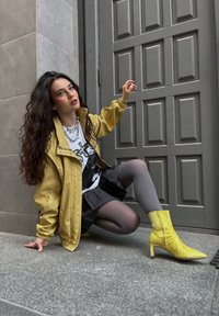Gelbe, gesteppte Knöchelboots mit spitzen Zehen und einem klobigen Absatz, kombiniert mit einem grauen Rock und einer gelben Jacke über einem Grafik-T-Shirt.