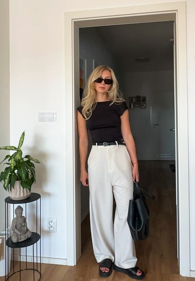 Top negro ajustado, pantalones de pierna ancha crema, sandalias planas negras, bolso negro oversized y gafas de sol oscuras. Entorno interior con plantas.