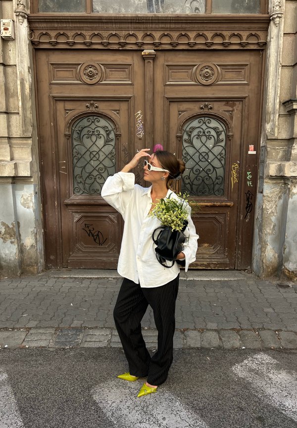 Femme en chemise blanche, pantalon noir et chaussures jaunes, tenant un sac noir avec des fleurs, debout dans la rue devant des portes en bois marron ornées.