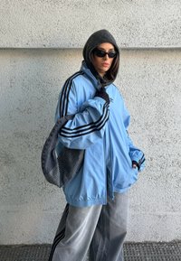 Femme portant une capuche noire, des lunettes de soleil, une veste bleu clair surdimensionnée avec des rayures noires, un pantalon gris, et portant un sac noir devant un mur texturé.