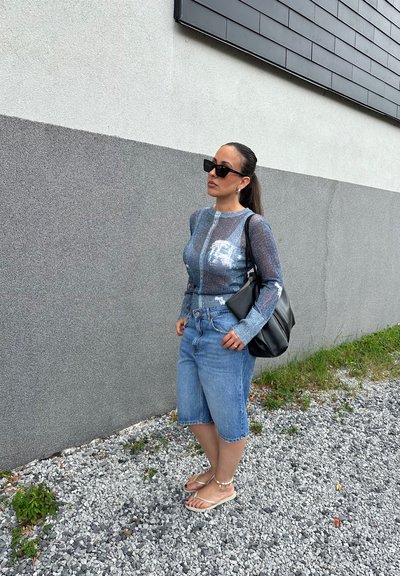 Blusa larga de manga larga, ligera y azul claro con un gráfico, combinada con pantalones cortos bermuda azul claro. Bolso negro y chanclas con detalles de cuentas.