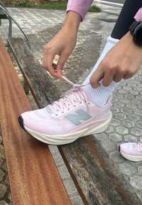 Lys rosa sportssko med mesh-overdel, grå detaljer og hvit såle, som blir knyttet av en hånd over en benk i tre.