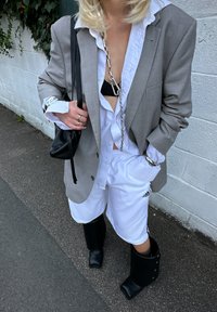 Šedý blazer přes bílou košili, černý bikiny top a bílé šortky s logem Adidas. Černé kožené vysoké boty až ke kolenům, stříbrný řetězový detail.